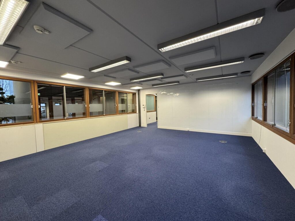 Empty office space with blue carpet, white walls, and large windows at De Ruijterkade.